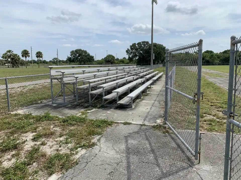 Illustration of the visiting side of a high school football field with empty aluminum bleachers and an open chain-link gate