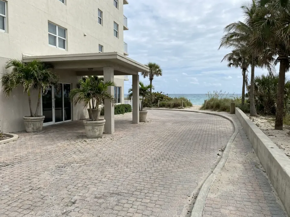 Illustration of a short curved hotel driveway with a small entrance canopy and potted palms, the beach and ocean past the end of the drive