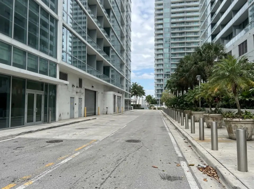 Illustration of a quiet Brickell service drive between glass towers, with bollards, planters and palms beside an empty loading area