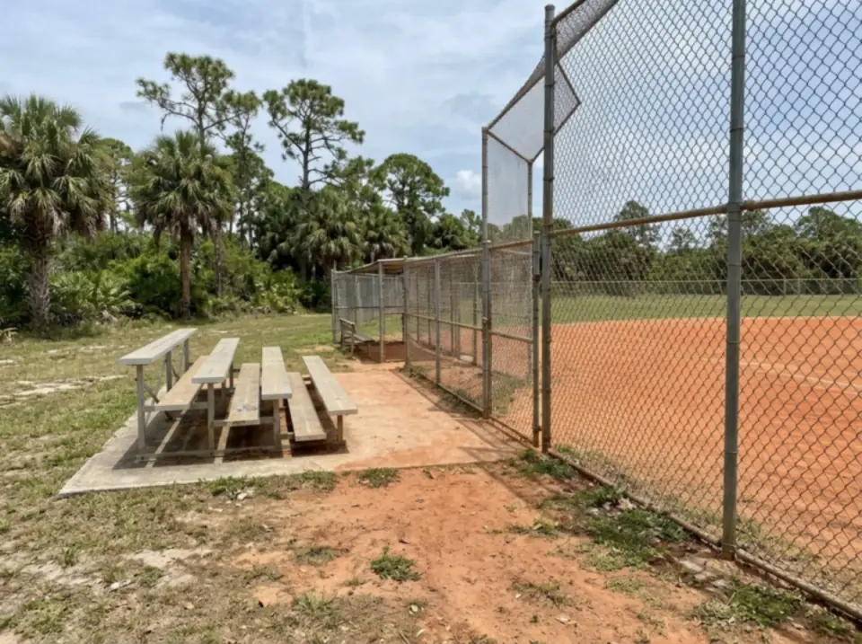 Illustration of an empty community baseball diamond in Port St. Lucie with aluminum bleachers, a chain-link backstop and palms and pines beyond the fence