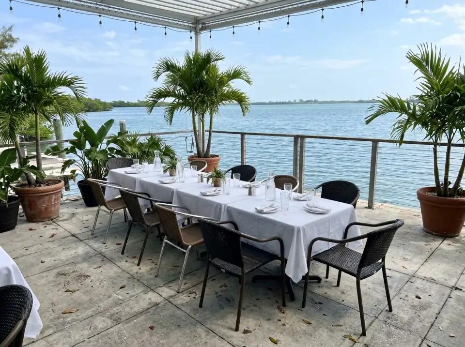Illustration of a waterfront restaurant terrace set for a group lunch, long white-clothed tables, potted palms and open water beyond the railing