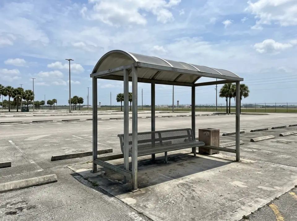 Illustration of an empty covered shuttle shelter with a metal bench at the edge of a very large, empty parking lot near an airport, light poles and palms