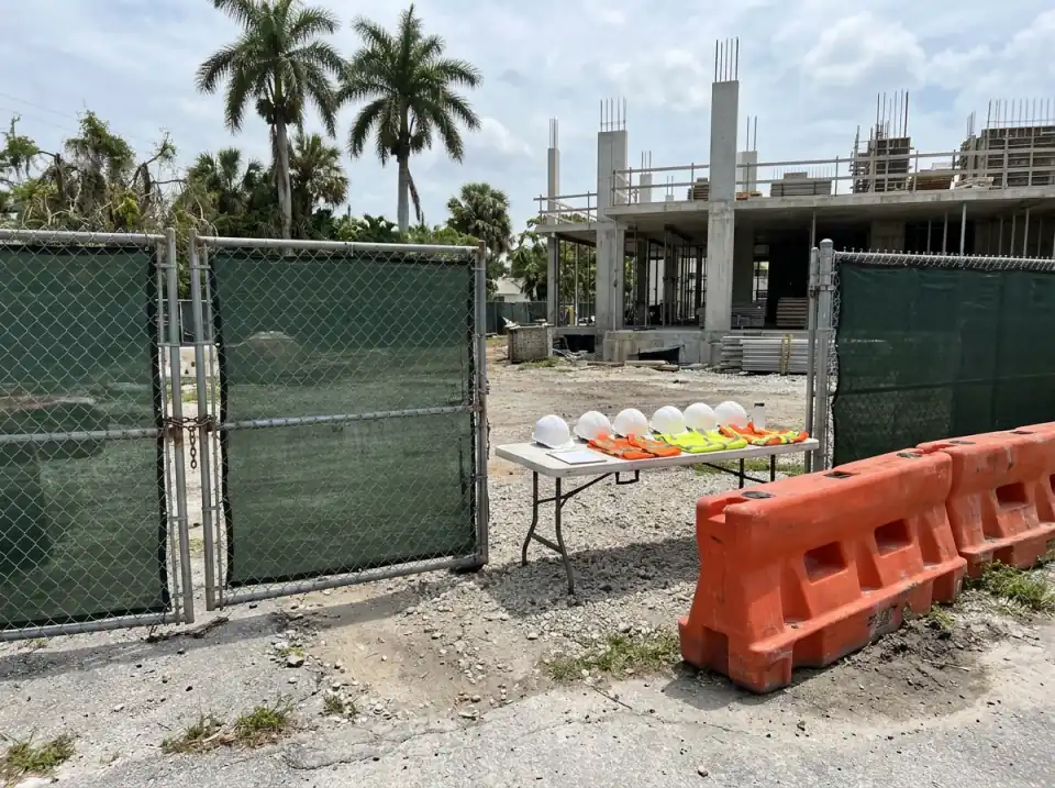 Illustration of a construction site entrance gate with green mesh fencing, orange barriers and a folding table holding white hard hats and safety vests