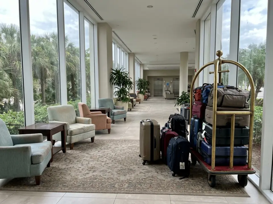 Illustration of a long hotel lobby with a brass bell cart stacked with suitcases beside armchairs and tall windows onto palms