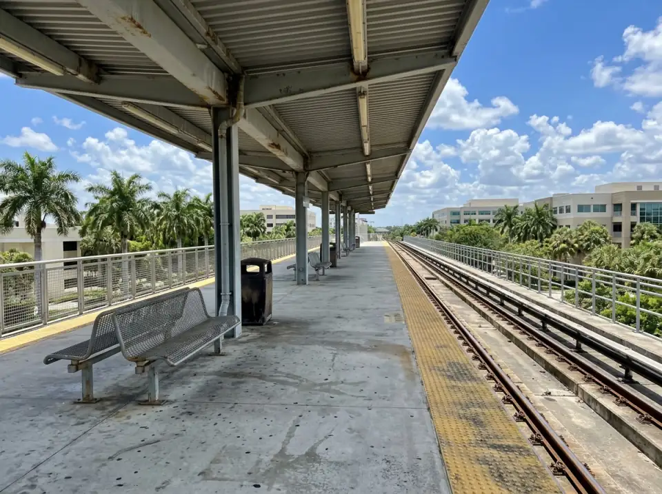Illustration of an empty elevated commuter rail platform under a long metal canopy, benches, palms and low office buildings beyond