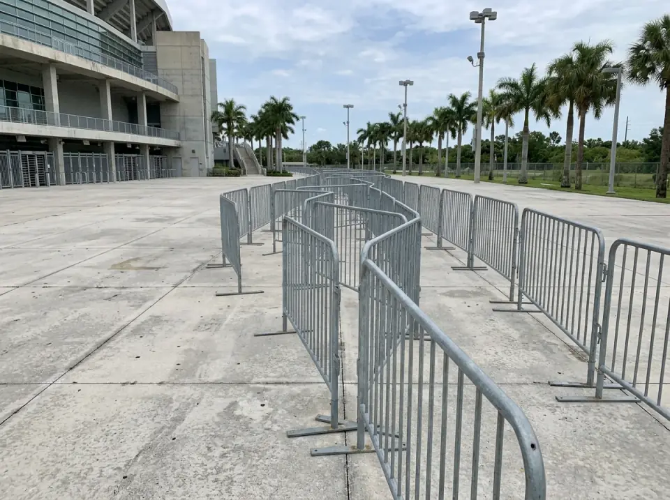 Illustration of a long empty queue lane of metal crowd barriers on a concrete plaza outside a venue, palms and light poles beyond