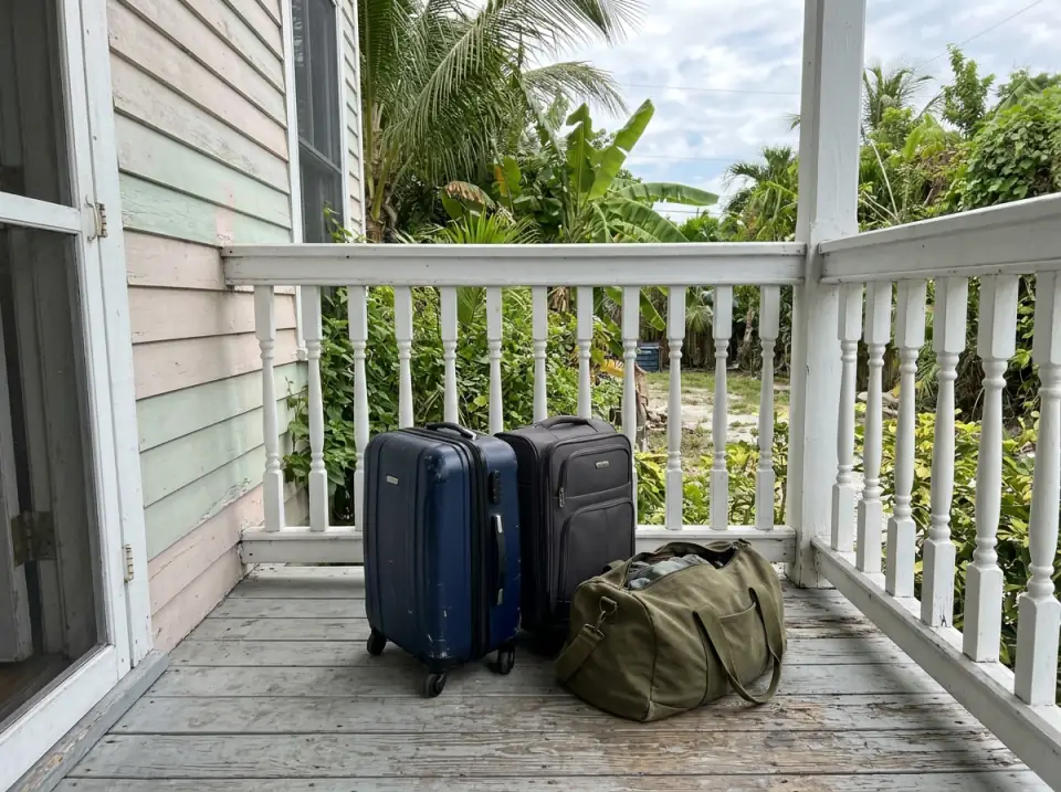 Illustration of small suitcases and a duffel bag on the wooden porch of a Key West guesthouse with a white railing and palms
