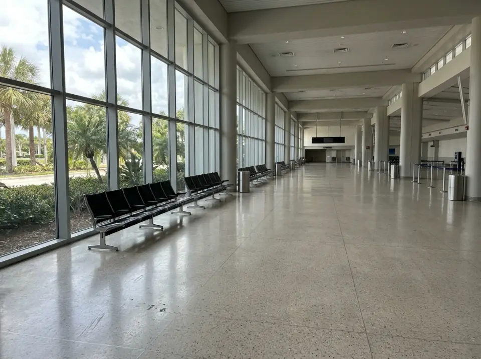 Illustration of a bright, empty airport arrivals hall with tall glass walls, palms outside and rows of black seats along the windows