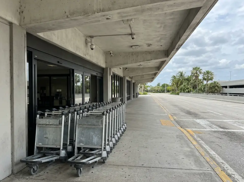 Illustration of steel luggage carts lined up outside an airport departures entrance under a concrete canopy, palms beyond the empty roadway