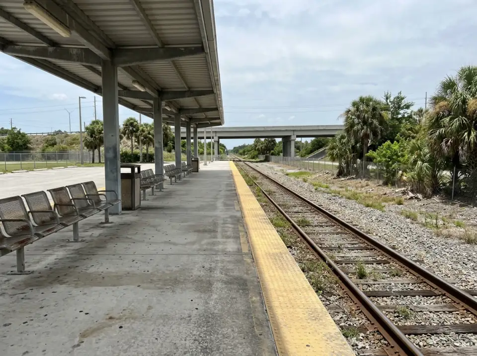 Illustration of an empty commuter rail platform under a steel canopy, empty tracks alongside, palms and an overpass in the distance