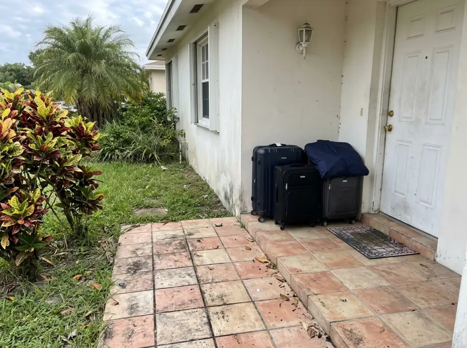 Illustration of packed suitcases and a garment bag waiting on the tiled front porch of a South Florida home, crotons and a palm in the yard