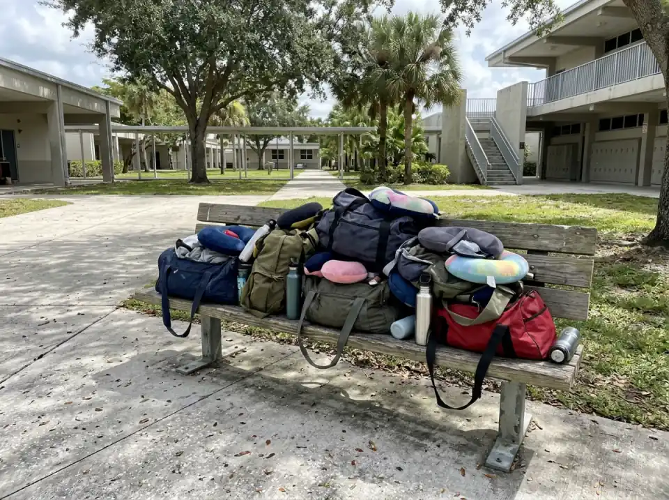 Illustration of duffel bags, travel pillows and water bottles piled on a wooden bench in a South Florida high school courtyard under shade trees