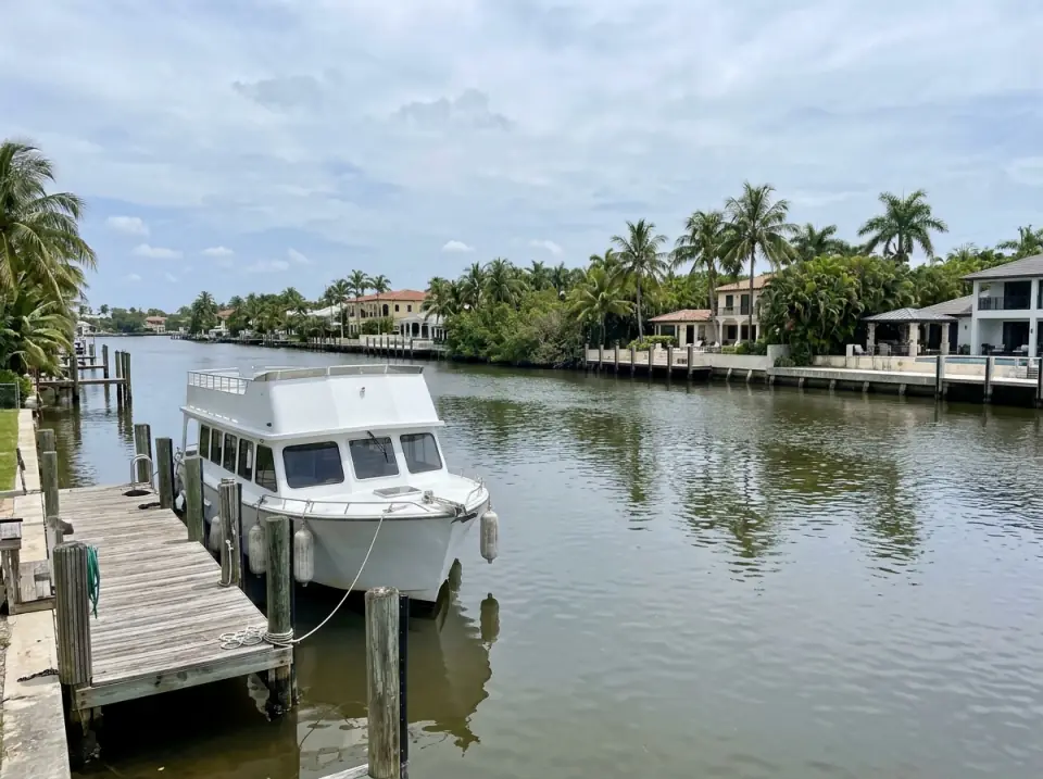 Illustration of a wide calm river in Fort Lauderdale lined with palms, waterfront homes and private docks, a plain white tour boat tied at a wooden landing
