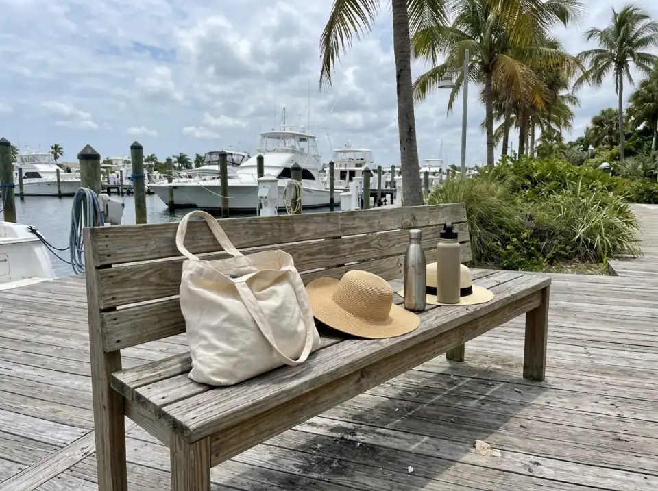 Illustration of straw sun hats, water bottles and a canvas tote bag on a wooden bench beside a marina boardwalk, moored white boats behind