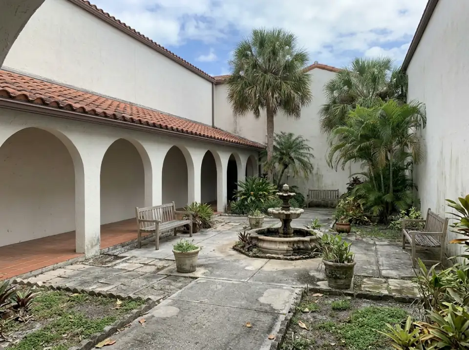 Illustration of a quiet church courtyard with an arched covered walkway, wooden benches, a small fountain and palms