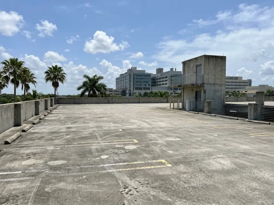 Illustration of the open top deck of an empty multi-level parking garage, painted parking lines, a stair tower and palms, a modern hospital campus building beyond
