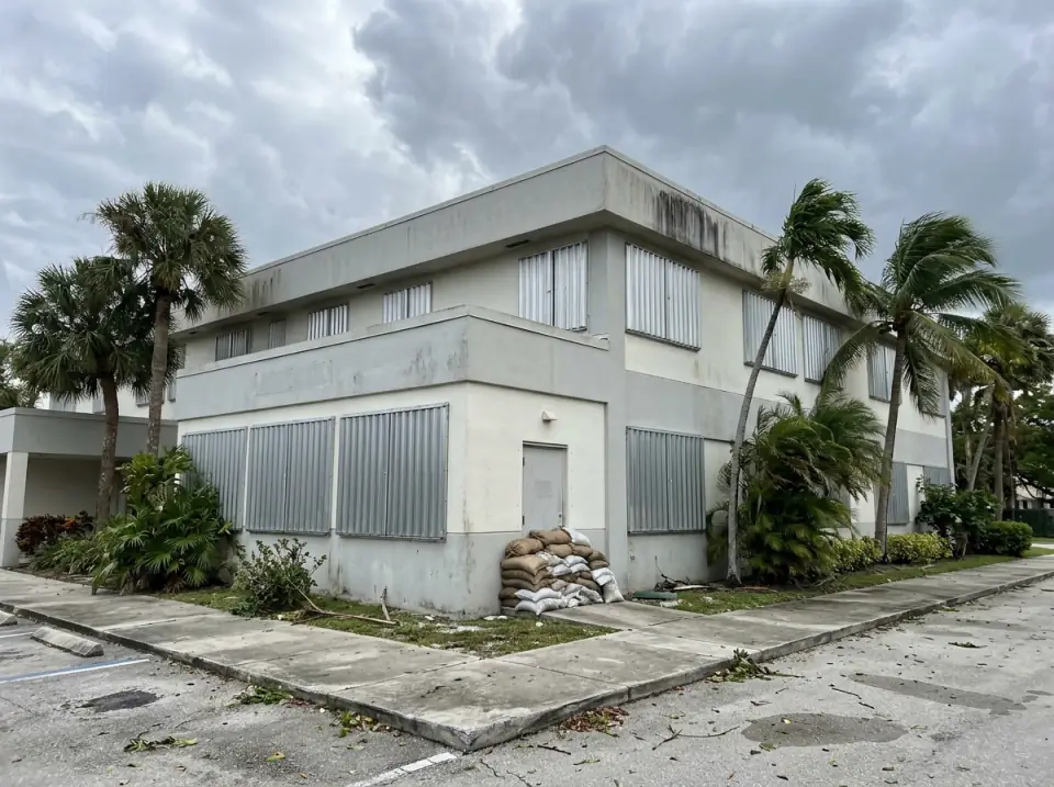 Illustration of metal storm shutters closed over the windows of a low hospital building, sandbags stacked by a side door and palms bending under heavy grey clouds