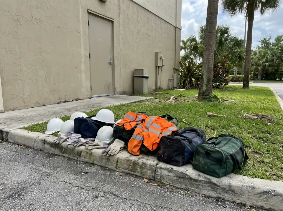 Illustration of white hard hats, work gloves, orange safety vests and duffel bags set on a curb outside a plain hotel side entrance with palms