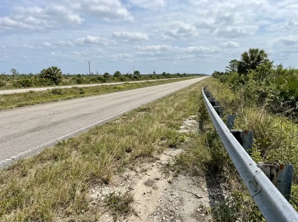 Illustration of an empty divided highway running straight through flat open South Florida country, seen from a roadside pull-off with a guardrail