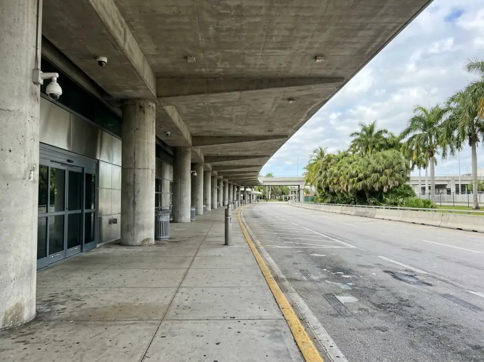 Illustration of an empty airport departures curb under a long concrete canopy, glass doors on one side and palms beyond the roadway