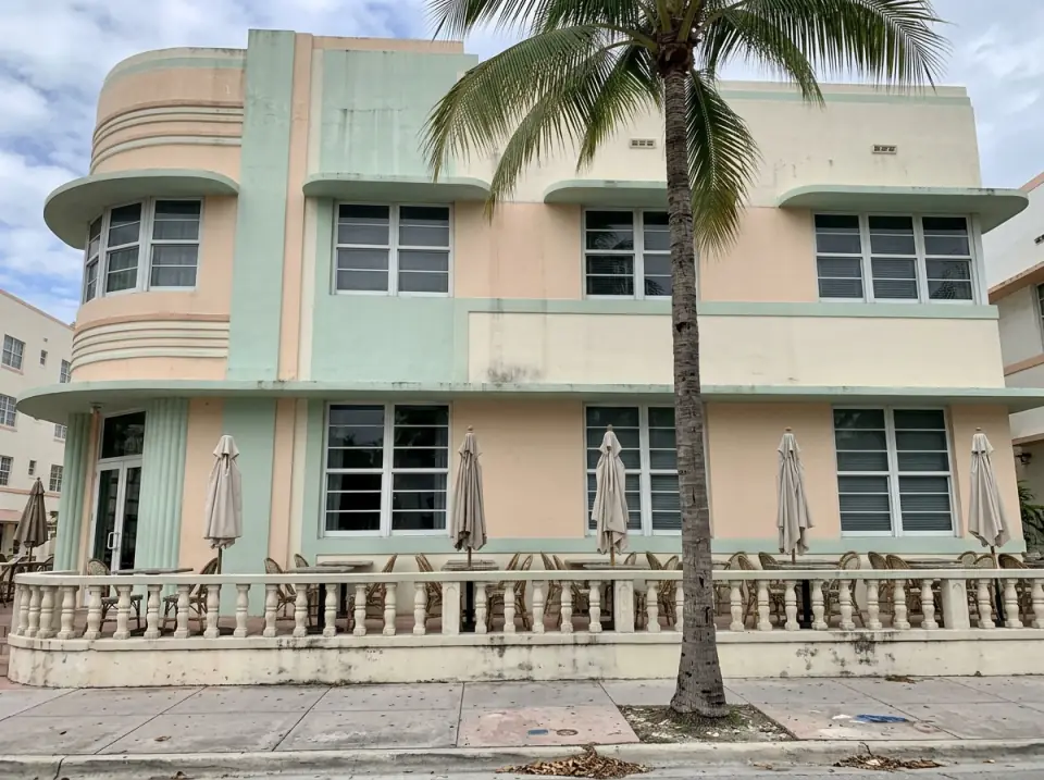 Illustration of a pastel Art Deco style hotel facade in South Beach with rounded corners, a porch of empty tables and a palm in front