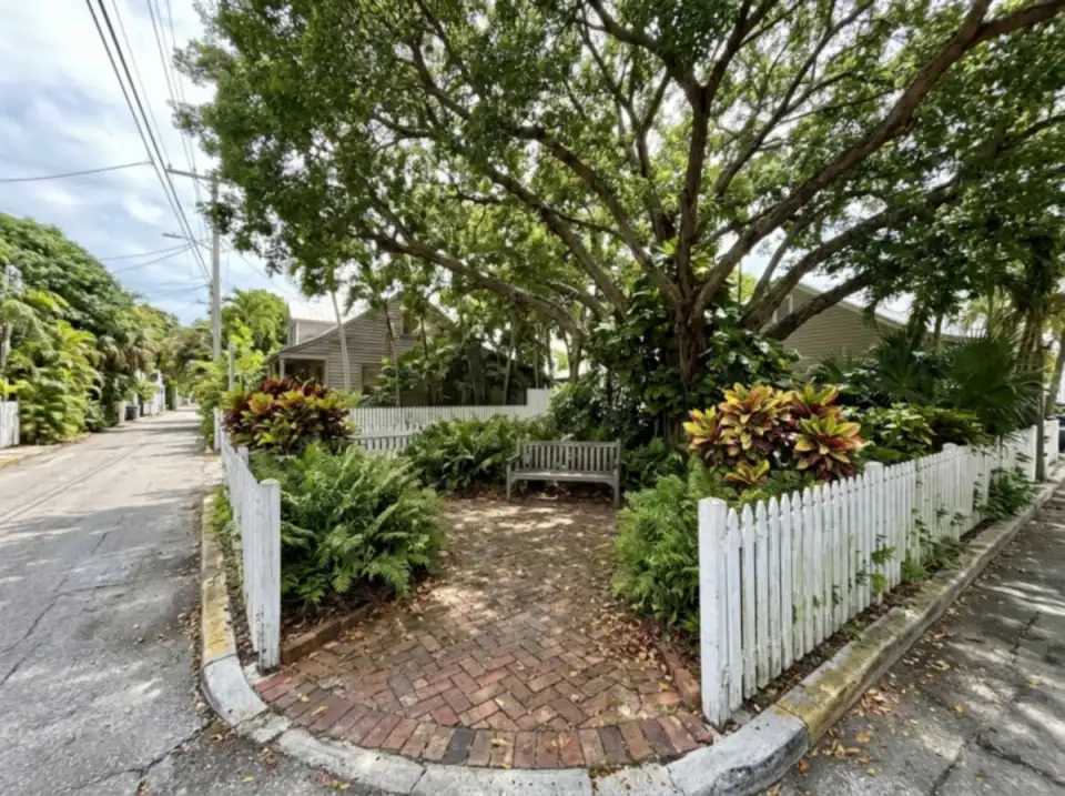 Illustration of a small shaded pocket park at a street corner in Old Town Key West, a bench under a big tree and a white picket fence