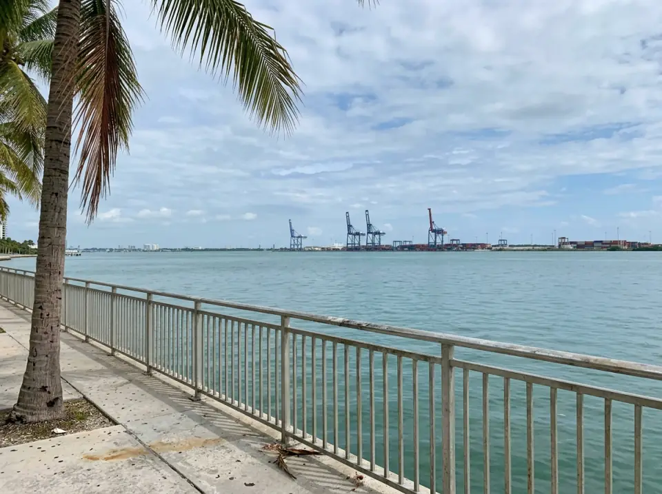 Illustration of Biscayne Bay seen from a downtown Miami waterfront walkway, the port's cargo cranes across the water