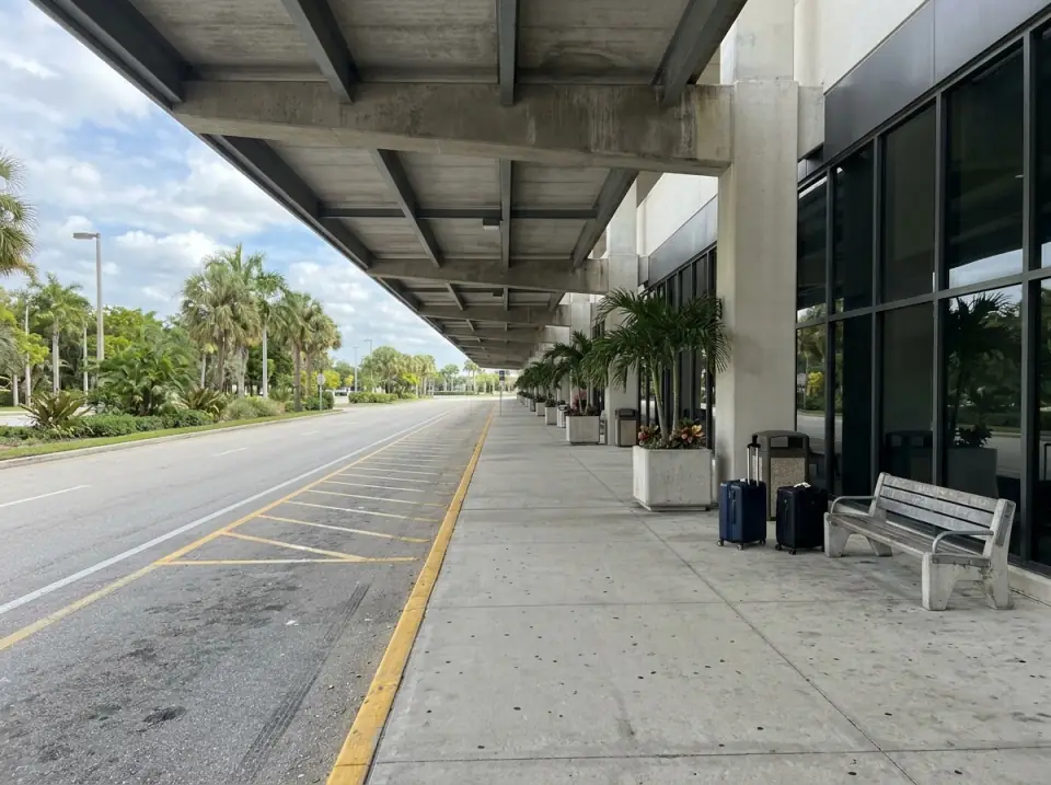 Illustration of an empty airport arrivals curb in West Palm Beach under a long canopy, two suitcases by a bench and palms in planters