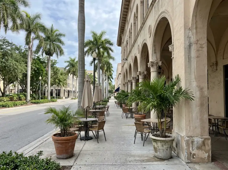 Illustration of empty cafe tables under the arched arcade of a Mediterranean revival building on a palm-lined downtown West Palm Beach street