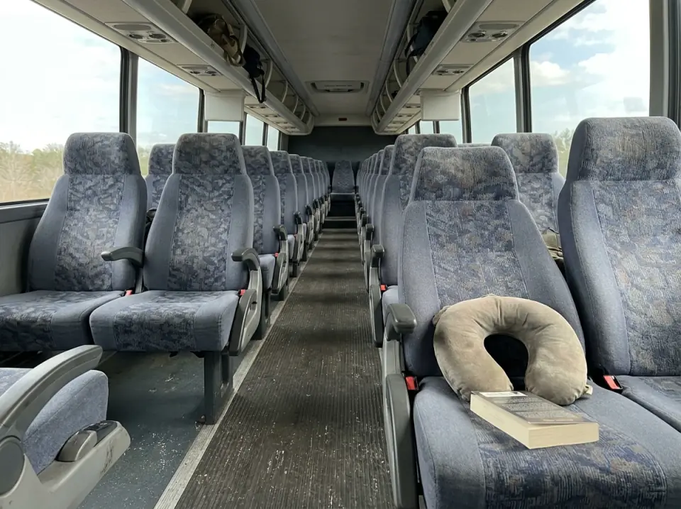 Illustration of the aisle of an empty motorcoach, a neck pillow and a paperback on a reclining seat and small backpacks in the overhead rack
