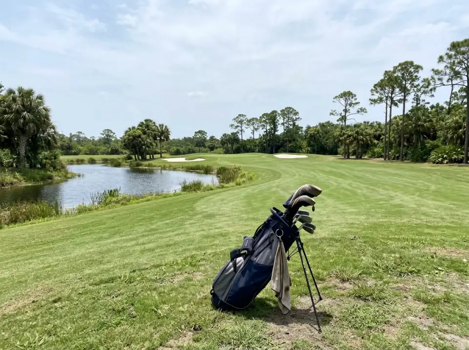 Illustration of an empty golf fairway bending around a small lake with sand bunkers and palms, a golf bag on its stand in the foreground