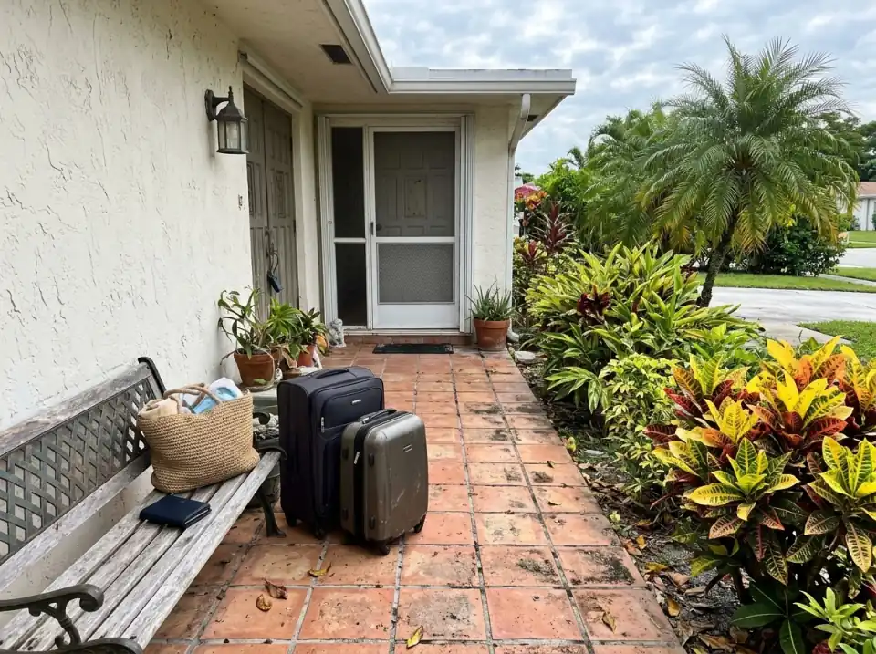 Illustration of two suitcases, a straw beach bag and a passport wallet waiting on a bench beside the front door of a South Florida home, crotons and a palm in the yard