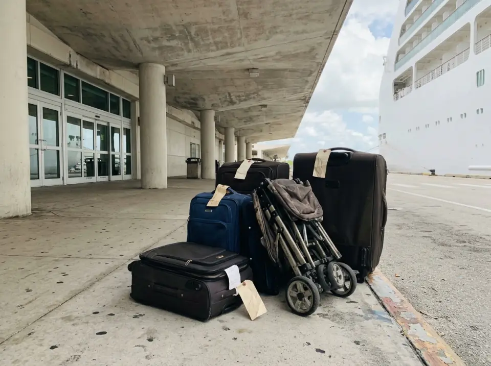 Illustration of suitcases with blank tags and a folded stroller on the sidewalk outside a cruise terminal exit, a plain white ship at the edge of the frame
