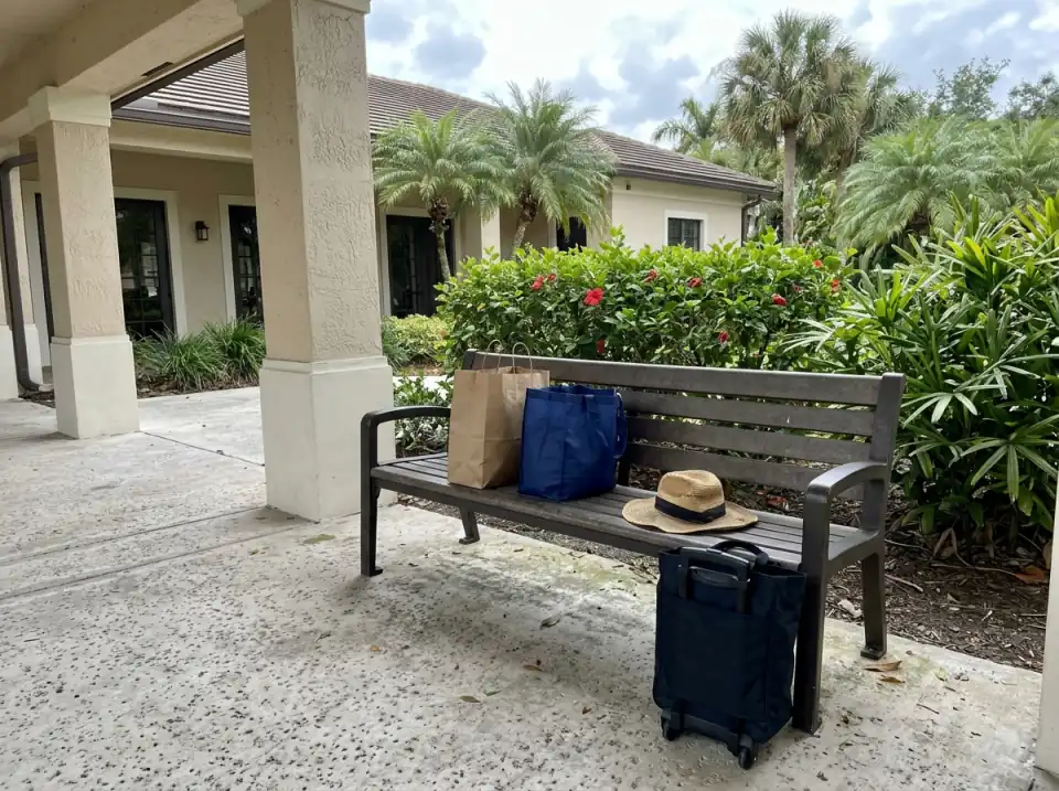 Illustration of shopping bags, a small rolling tote and a straw sun hat on a bench beside a covered walkway outside a community clubhouse