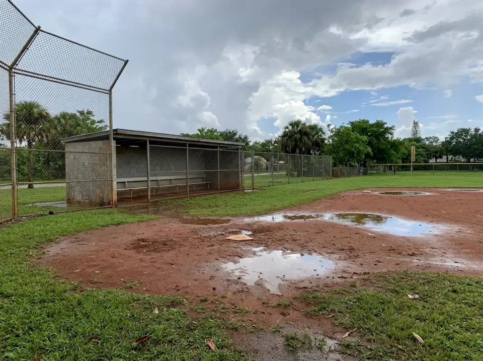 Illustration of an empty softball field after a summer rain shower, puddles on the infield, a dugout and chain-link fence under breaking clouds