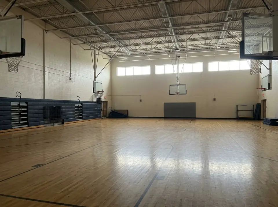 Illustration of an empty indoor gym with a polished basketball court, folded bleachers along one wall and high windows
