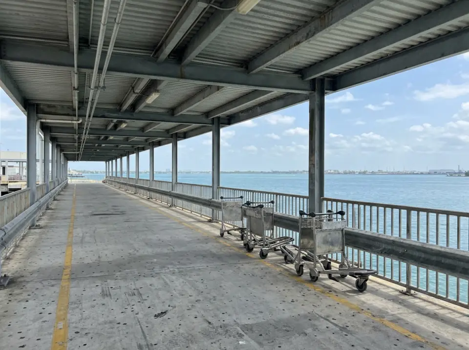 Illustration of an empty covered walkway at a Miami cruise port, luggage carts along the railing and Biscayne Bay beyond
