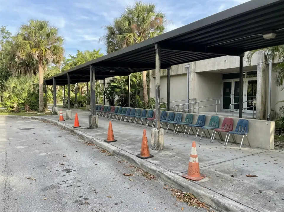 Illustration of a shaded camp pickup area beside a recreation center, a long covered walkway with a row of empty plastic chairs and orange cones on the curb