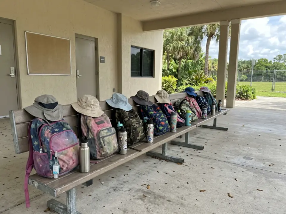 Illustration of kids' backpacks, refillable water bottles and sun hats lined up on a long bench under a covered walkway outside a community recreation center