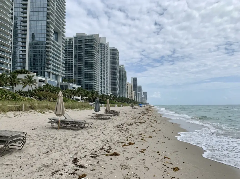 Illustration of the beach at Sunny Isles Beach looking north, with a long row of tall glass towers behind the sand and closed umbrellas