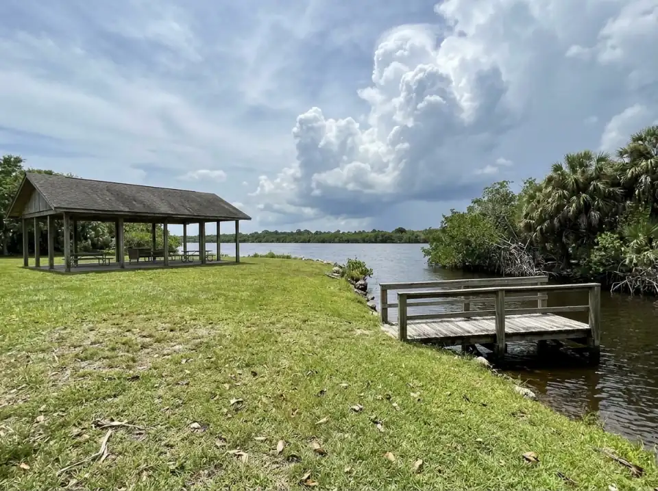 Illustration of a riverside lawn in Port St. Lucie with an open pavilion and a small wooden dock, mangroves on the bank and storm clouds building over the far shore