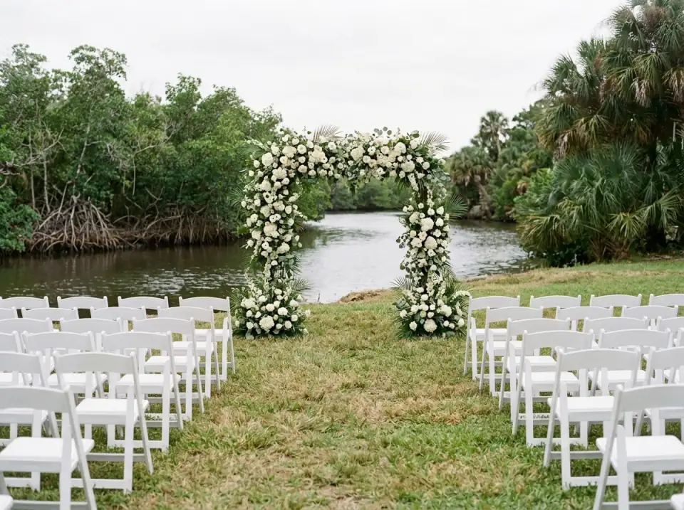 Illustration of a wedding arch of white flowers on a lawn beside a calm river with mangroves, rows of empty white chairs facing it