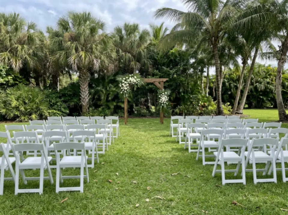 Illustration of rows of white folding chairs on a lawn facing a wooden arbor with white flowers, palms around the ceremony site